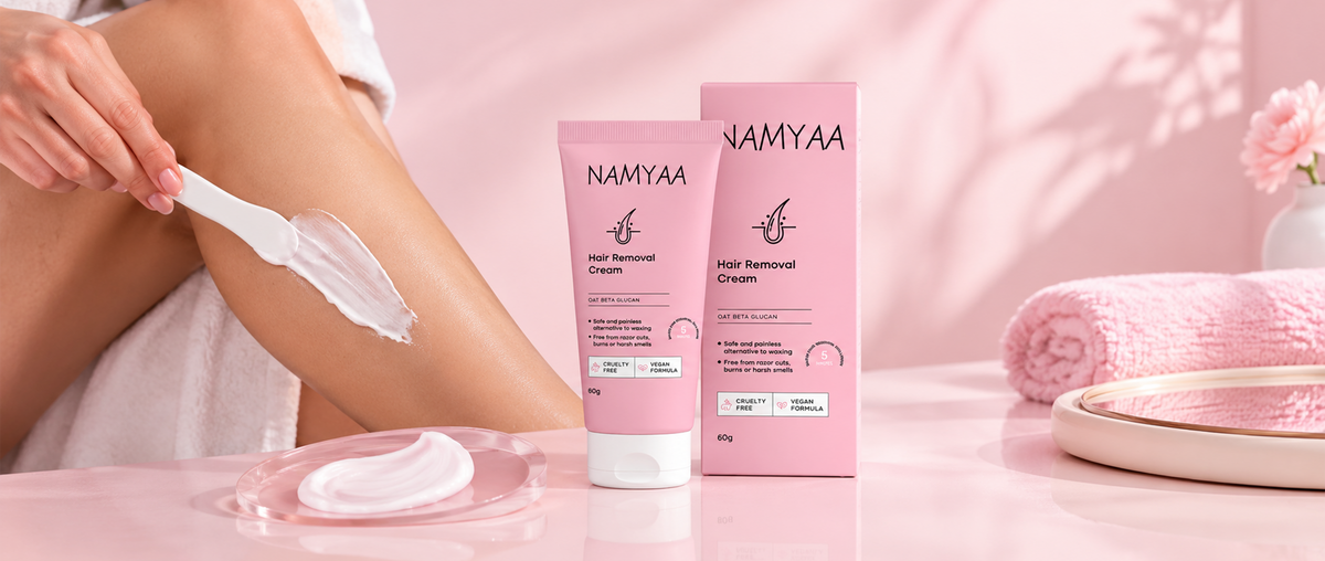 A close-up of a woman applying white hair removal cream to her lower leg using a spatula, next to a tube and box of Namyaa Hair Removal Cream on a pink table.