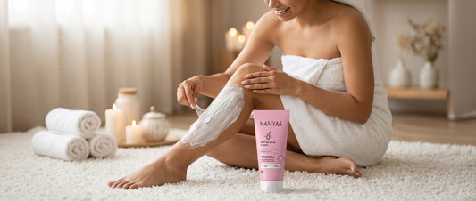 A woman wrapped in a white bath towel sitting on a plush rug, using a plastic spatula to apply Namyaa Hair Removal Cream to her leg next to burning candles and rolled towels
