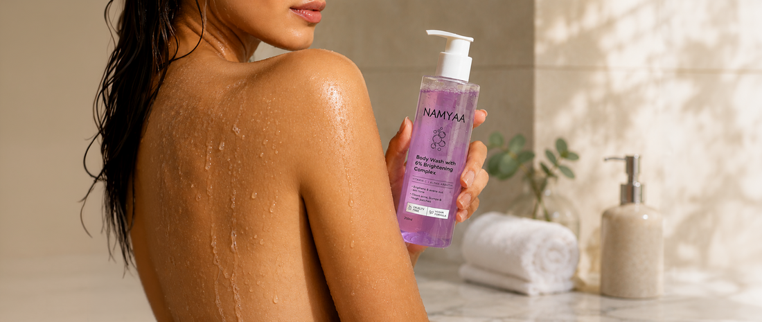 A close-up of a woman with wet, glowing skin holding a purple bottle of Namyaa Body Wash with 6% Brightening Complex in a sunlit bathroom setup with rolled white towels.
