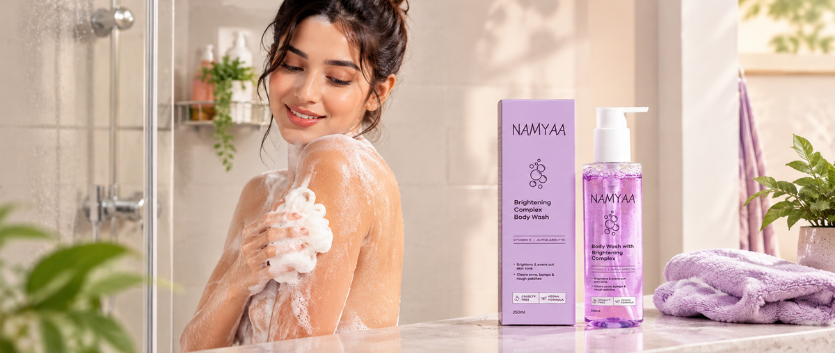 A smiling woman in the shower using a white loofah with lather on her shoulder, with a pump bottle and box of purple Namyaa Brightening Complex Body Wash in the foreground.