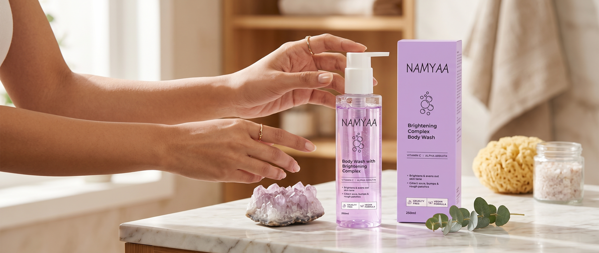A person pressing the pump of a clear bottle of purple Namyaa Brightening Complex Body Wash next to its matching outer box on a marble counter.