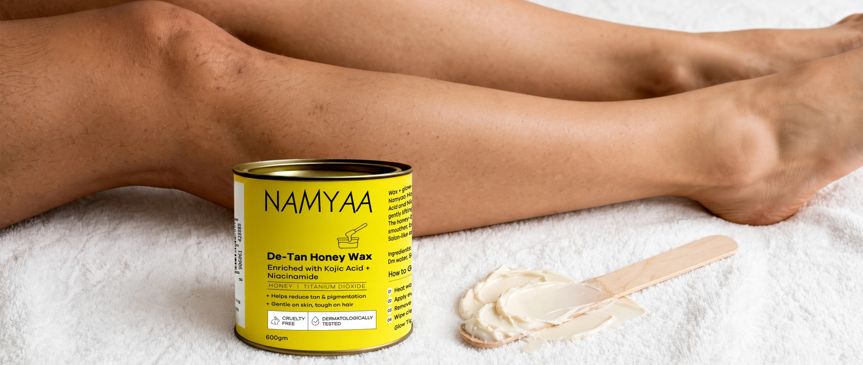 A yellow tin of Namyaa De-Tan Honey Wax next to a wooden spatula covered in cream-colored wax, placed in front of a pair of legs resting on a white towel.