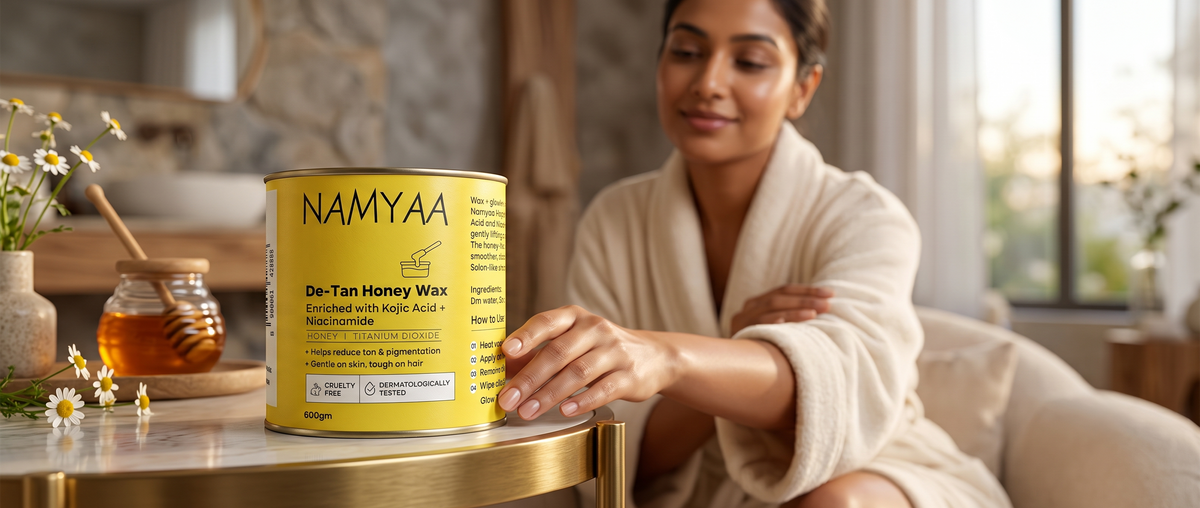 A woman in a bathrobe reaching toward a yellow tin of Namyaa De-Tan Honey Wax set on a table next to a pot of honey and chamomile flowers.