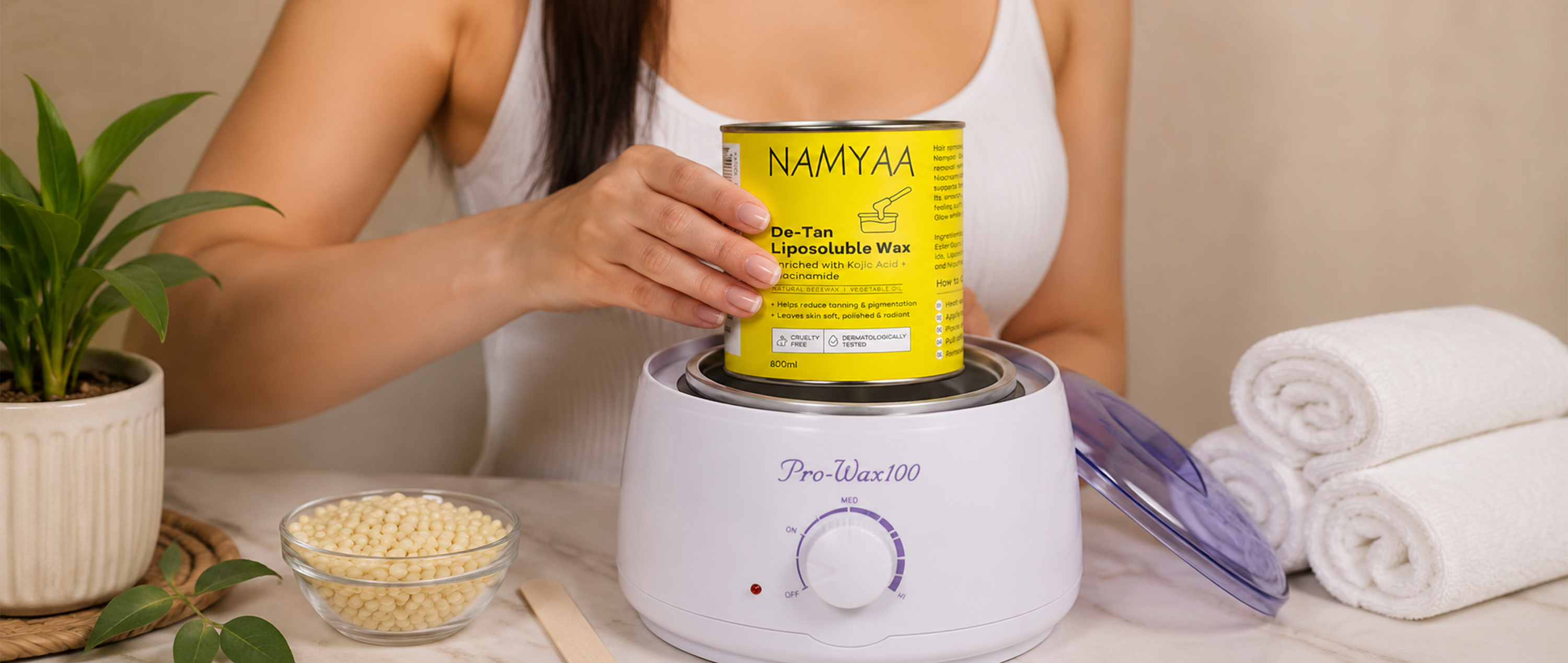 A woman placing a yellow tin of Namyaa De-Tan Liposoluble Wax into a white Pro-Wax 100 electric wax heater next to a bowl of wax beans and rolled towels.