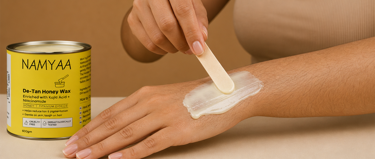 A close-up of a person using a wooden spatula to apply a smooth layer of cream-colored wax onto their arm, next to a yellow tin of Namyaa De-Tan Honey Wax enriched with Kojic Acid and Niacinamide.