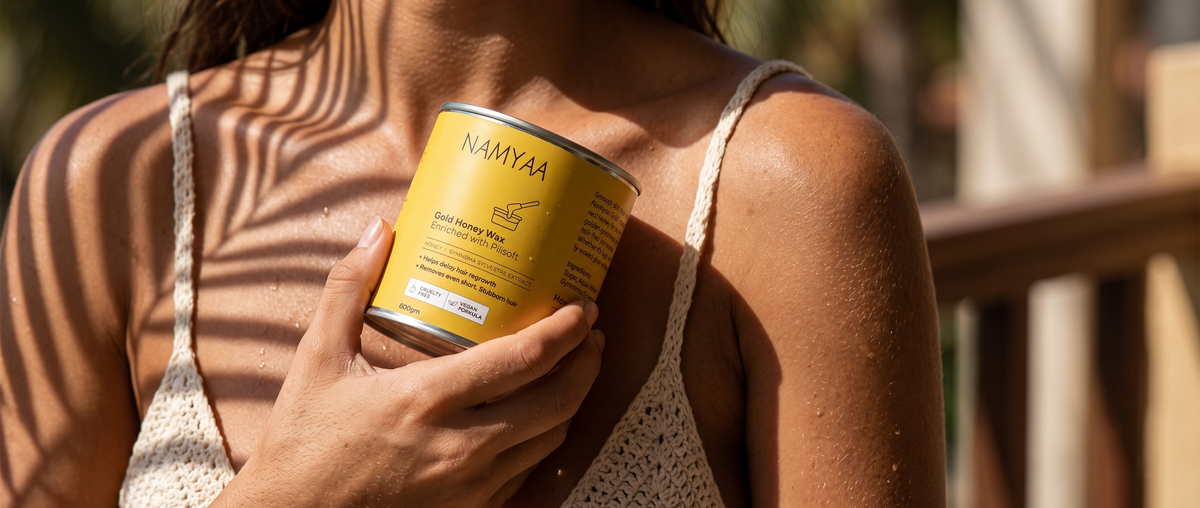 A woman holding a yellow tin of Namyaa Gold Honey Wax against her chest under palm leaf shadows in warm sunlight.