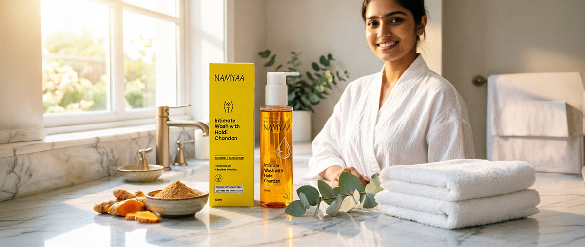 A South Asian woman in a white robe smiling next to Namyaa Haldi Chandan intimate wash on a sunlit marble bathroom counter with fresh eucalyptus and towels.