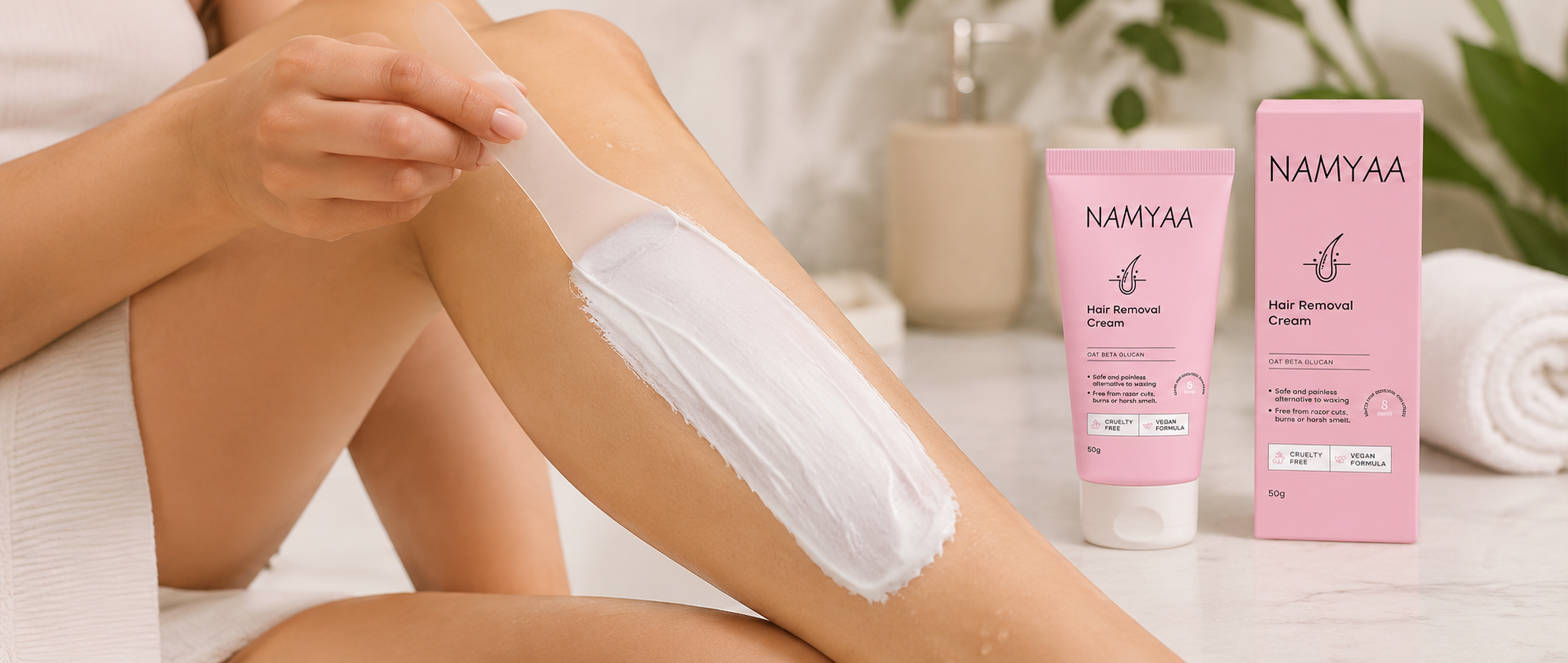 Close-up of a person using a translucent spatula to spread white hair removal cream onto their leg, next to a pink tube and outer box of Namyaa Hair Removal Cream with Oat Beta Glucan.