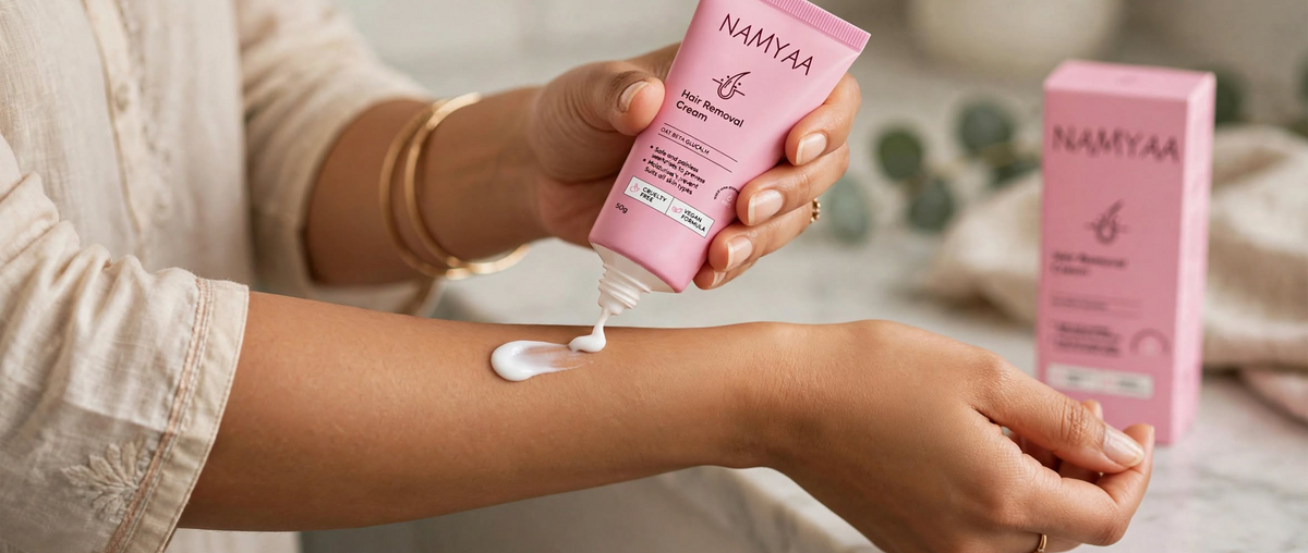 A close-up of a person squeezing white cream from a pink Namyaa Hair Removal Cream tube onto their forearm, with the product's outer box in the blurred background.
