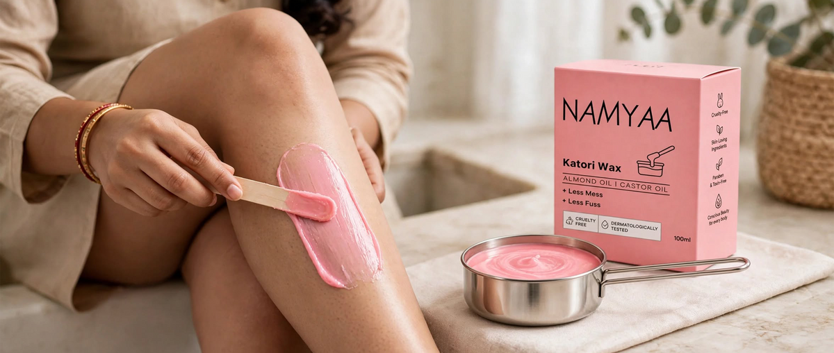 A woman applying smooth pink wax onto her lower leg using a wooden spatula, next to a small metal wax pot and the Namyaa Katori Wax pink box.