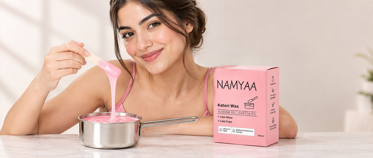A smiling woman lifting dripping pink wax with a wooden spatula from a metal pot next to the Namyaa Katori Wax pink box.