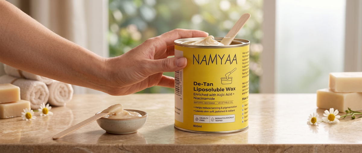 A hand reaching for a yellow tin of Namyaa De-Tan Liposoluble Wax set on a marble table with a wooden spatula in a small wax bowl beside it.