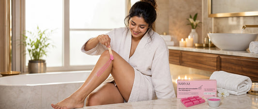 A smiling woman in a white waffle bathrobe applying pink wax to her leg using a wooden spatula, next to the Namyaa Brazilian Microwaveable Hot Waxing Kit on a marble bathroom counter.