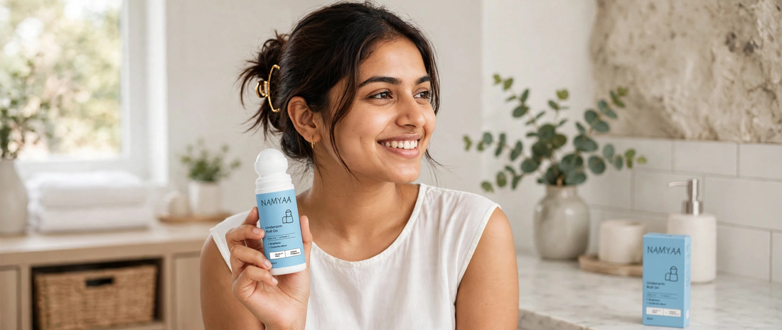 A smiling young woman holding a Namyaa Underarm Roll-On deodorant bottle in a bright bathroom setting, with the product packaging visible on the counter behind her.