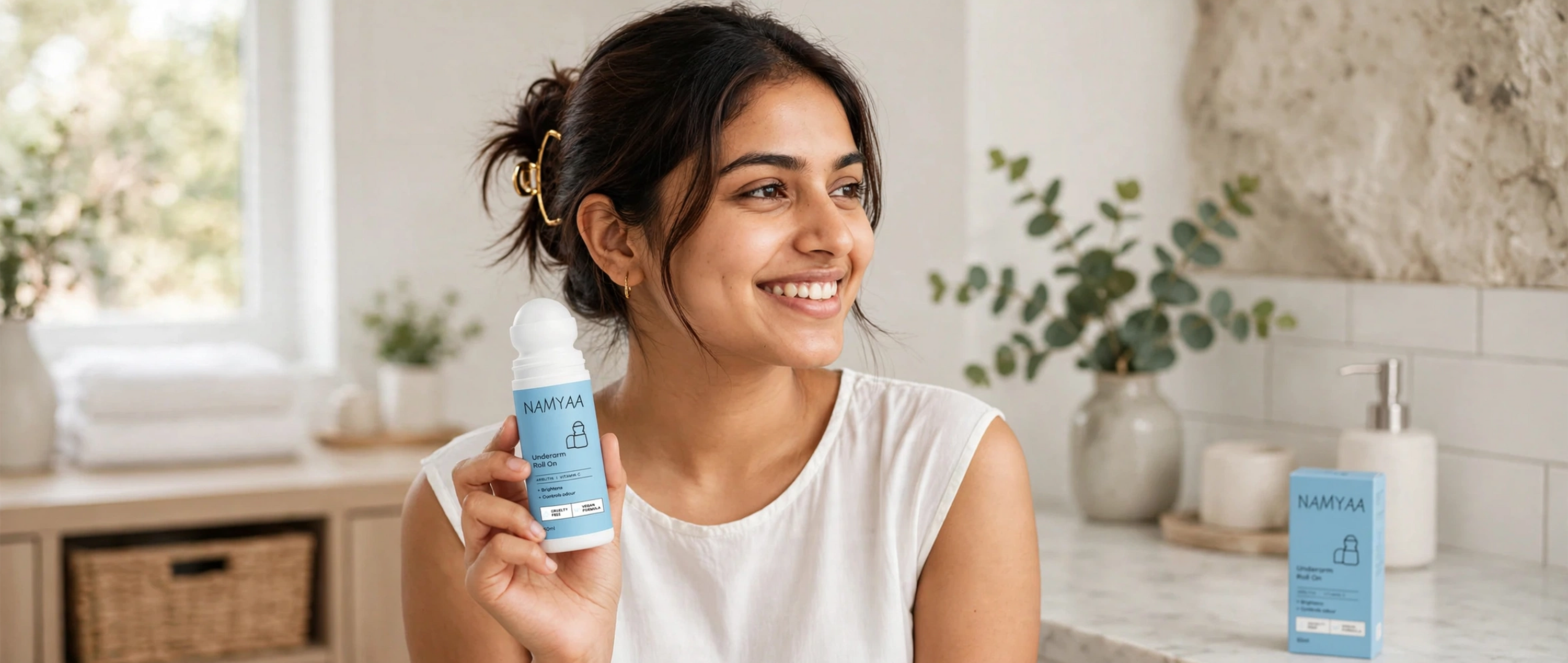 A smiling young woman holding a Namyaa Underarm Roll-On deodorant bottle in a bright bathroom setting, with the product packaging visible on the counter behind her.
