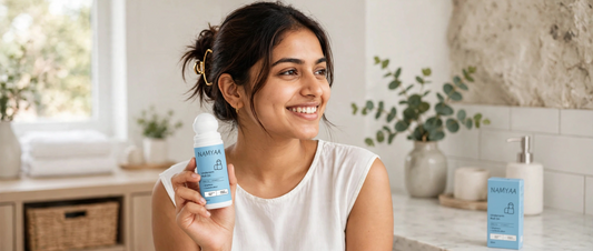 A smiling young woman holding a Namyaa Underarm Roll-On deodorant bottle in a bright bathroom setting, with the product packaging visible on the counter behind her.