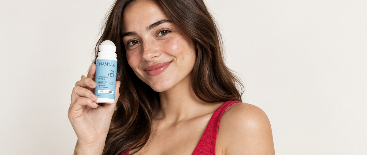 A smiling woman holding up a bottle of blue Namyaa Underarm Roll On deodorant next to her face against a neutral background.