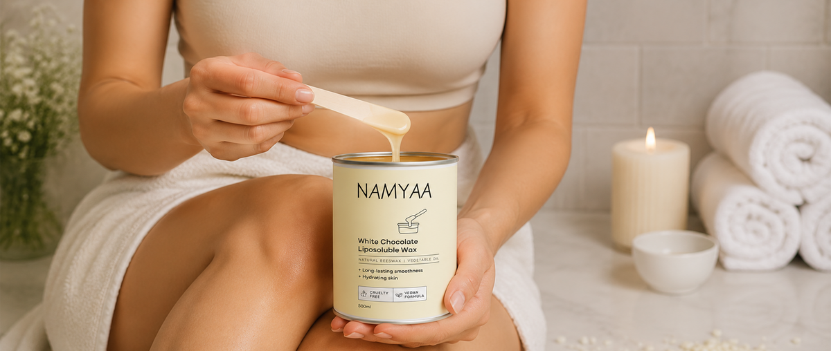 A person dipping a wooden spatula into a tin of Namyaa White Chocolate Liposoluble Wax in a spa-like bathroom setting with towels and a lit candle.