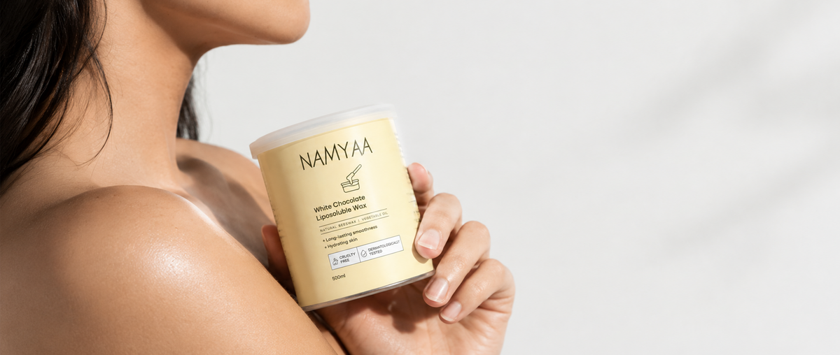 A close-up of a woman holding a tin of Namyaa White Chocolate Liposoluble Wax against her shoulder on a bright background.